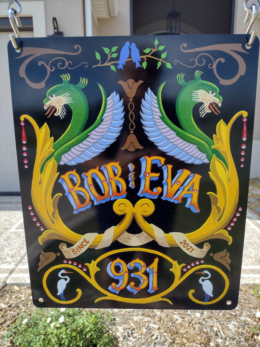 close up photo of the lamppost sign. It has 2 large acanthus scrolls with dragons emerging, several other flowers and birds, and the words "Bob & Eva" with the house number 931.